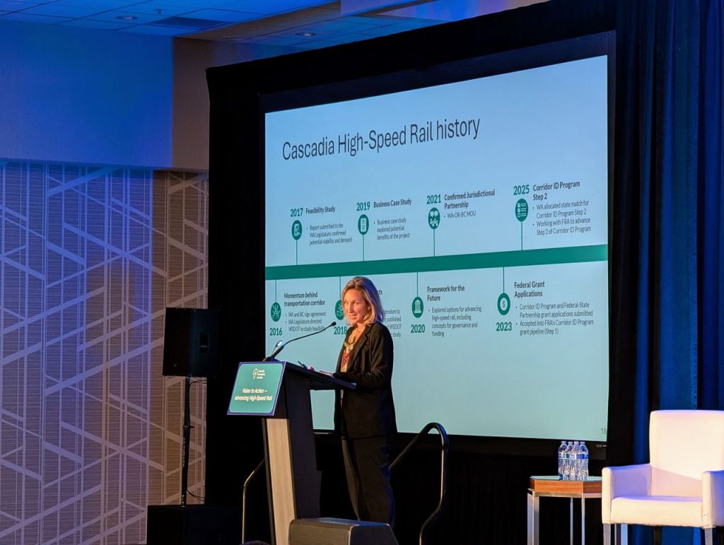 Levy is a blond woman standing at a lectern with the screen showing a timeline slide with milestones in Cascadia high speed rail planning.