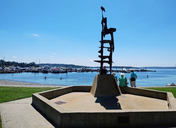 A tower-like metal sculpture crowns Kirkland's Marina Park, with a few pedestrians walking on the path along its edge. The marina and Moss Bay is in the distance.