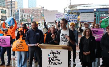 ‘Build the Damn Trains’ Campaign Pushes Sound Transit Toward Creative Solutions Advocates wave "build the damn trains sign" and Kirk from Transportation Choices Coalition speaks at the lectern