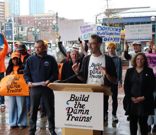 ‘Build the Damn Trains’ Campaign Pushes Sound Transit Toward Creative Solutions Advocates wave "build the damn trains sign" and Kirk from Transportation Choices Coalition speaks at the lectern