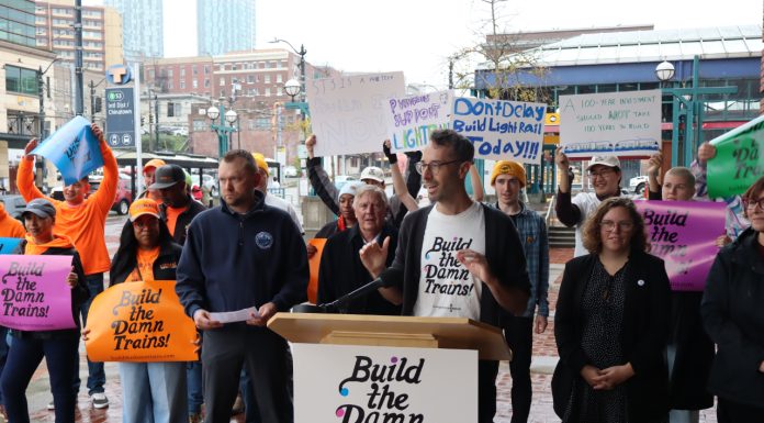 ‘Build the Damn Trains’ Campaign Pushes Sound Transit Toward Creative Solutions Advocates wave "build the damn trains sign" and Kirk from Transportation Choices Coalition speaks at the lectern