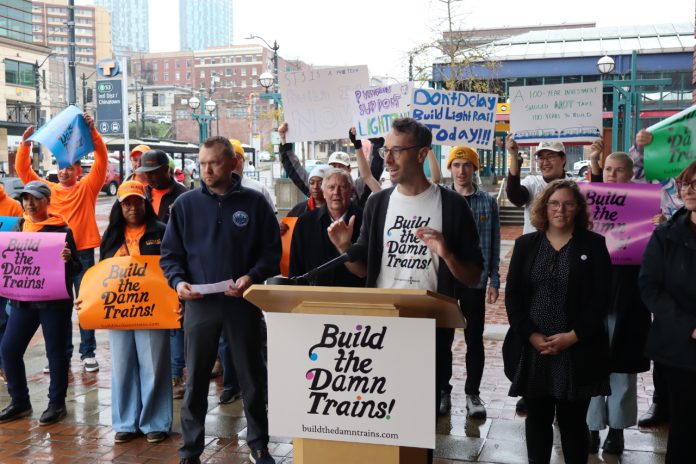 Build the Damn Trains press conference Advocates wave "build the damn trains sign" and Kirk from Transportation Choices Coalition speaks at the lectern