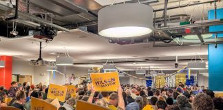 The view from the back of the room with supporters waving yellow Wilson signs and the candidate in the distance on stage.