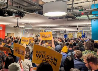 Wilson Trails in Early Results, Seattle Mayor’s Race on Nailbiter Course The view from the back of the room with supporters waving yellow Wilson signs and the candidate in the distance on stage.