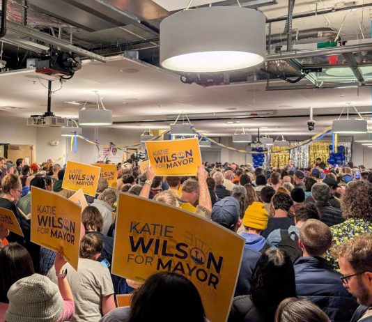 Wilson Trails in Early Results, Seattle Mayor’s Race on Nailbiter Course The view from the back of the room with supporters waving yellow Wilson signs and the candidate in the distance on stage.
