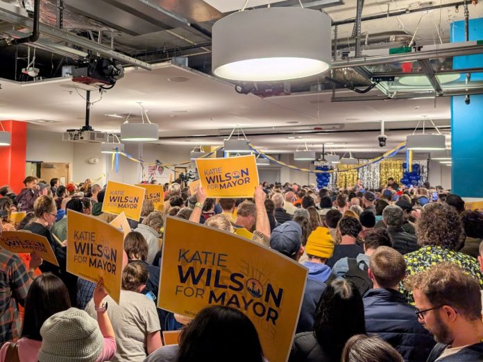 Katie Wilson election night party - The Urbanist 20251105 The view from the back of the room with supporters waving yellow Wilson signs and the candidate in the distance on stage.