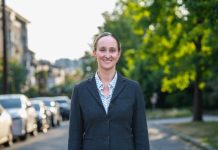 Op-Ed: Opioid Treatment Nurse Shares Why They Support Katie Wilson Wilson wears a blzer and stands on an urban street with trees and a line of parked cars in the background.