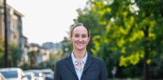 Op-Ed: Opioid Treatment Nurse Shares Why They Support Katie Wilson Wilson wears a blzer and stands on an urban street with trees and a line of parked cars in the background.