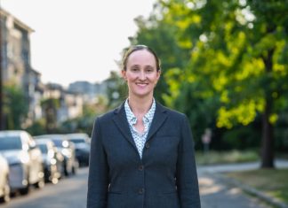 Katie Wilson Pulls Away from Harrell, Leads by 1,346 Votes Wilson wears a blzer and stands on an urban street with trees and a line of parked cars in the background.