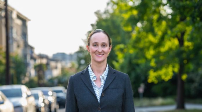 Op-Ed: Opioid Treatment Nurse Shares Why They Support Katie Wilson Wilson wears a blzer and stands on an urban street with trees and a line of parked cars in the background.