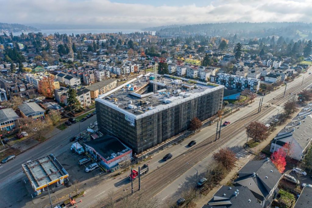 A yellow construction wrap is around the six-story building. Townhomes, treetops, and Lake Washington are in the background.