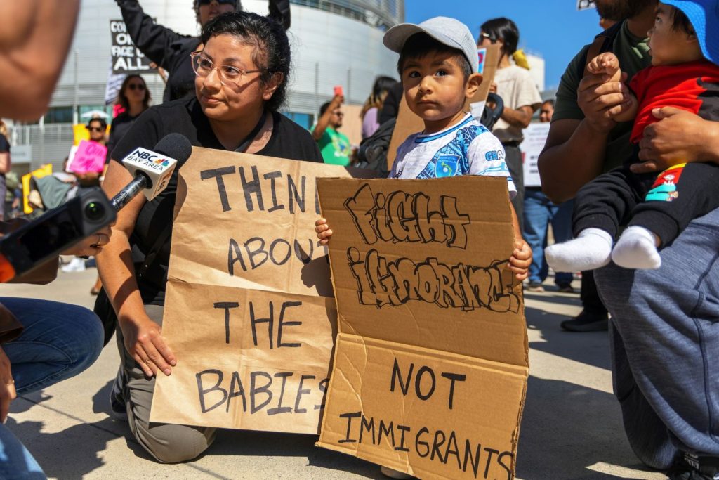 A Latina holds a sign saying "think about the babies." At her side, a young kid holds another cardboard sign saying "First ignorance, not immigrants" They are being interviewed by a NBC reporter. Behind them are more protesters.
