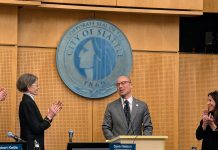 Eddie Lin Becomes Seattle’s Newest Councilmember Lin wears a gray suit and glasses and stands at the lectern with the City of Seattle seal behind him. Council President Sara Nelson and Maritza Rivera are to either side clapping.