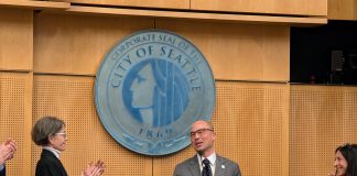 Eddie Lin Becomes Seattle’s Newest Councilmember Lin wears a gray suit and glasses and stands at the lectern with the City of Seattle seal behind him. Council President Sara Nelson and Maritza Rivera are to either side clapping.