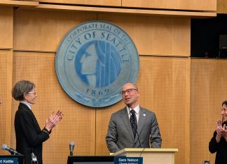 Eddie Lin Becomes Seattle’s Newest Councilmember Lin wears a gray suit and glasses and stands at the lectern with the City of Seattle seal behind him. Council President Sara Nelson and Maritza Rivera are to either side clapping.