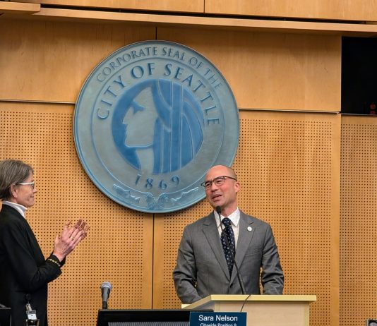 Eddie Lin Becomes Seattle’s Newest Councilmember Lin wears a gray suit and glasses and stands at the lectern with the City of Seattle seal behind him. Council President Sara Nelson and Maritza Rivera are to either side clapping.