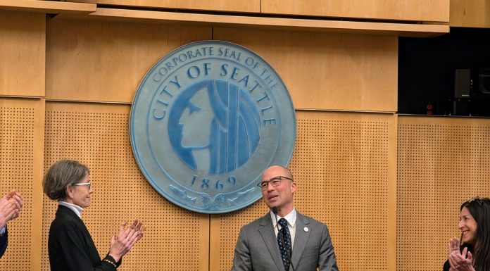Eddie Lin Becomes Seattle’s Newest Councilmember Lin wears a gray suit and glasses and stands at the lectern with the City of Seattle seal behind him. Council President Sara Nelson and Maritza Rivera are to either side clapping.