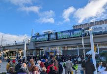 Urbanist Podcast: A Light Rail Opening and Police Accountability Door Closing A line of people approach Federal Way Downtown Station, with a train waiting at the elevated platform.