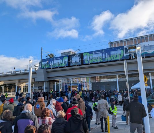 Urbanist Podcast: A Light Rail Opening and Police Accountability Door Closing A line of people approach Federal Way Downtown Station, with a train waiting at the elevated platform.