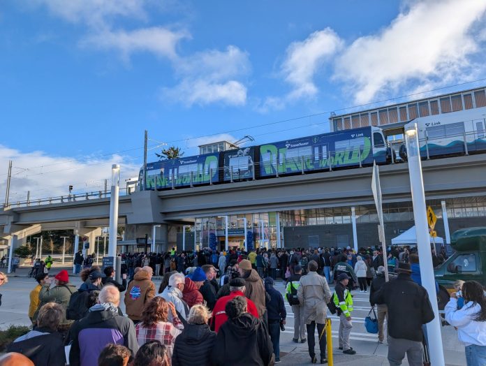 Federal Way Station ribboncutting crowd - Trumm 20251206 A line of people approach Federal Way Downtown Station, with a train waiting at the elevated platform.