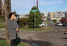 Despite Sidewalk Funding Boost, Some Seattle Neighborhoods Still Feel Shortchanged Dana is an Asian woman with black hair wearing a gray coat. She stands on a sidewalk-less corner by a utility pole. In the distance are some large apartment complexes.