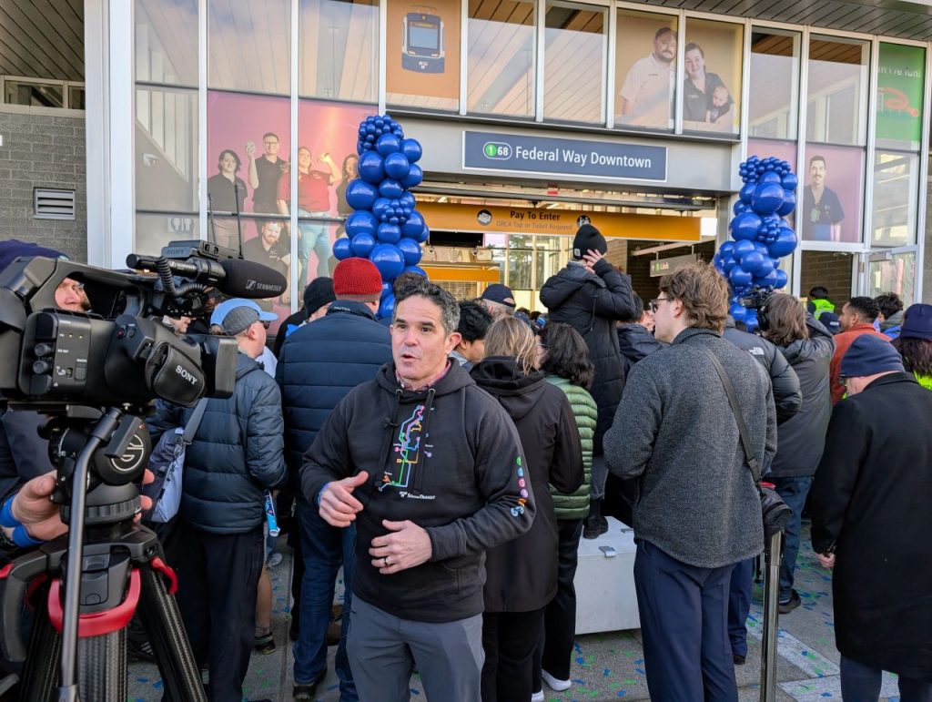 Mello stands in front of a TV camera giving an interview just at officials cut the ribbon on Federal Way's new light rail station. Pillars of blue balloons adorn the station entrance.