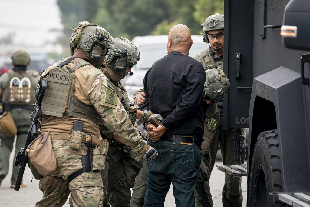 An image of I.C.E. "enforcement and removal operation" in Los Angeles provided by the Department of Homeland Security shows three large agents in camo and battle helmets handcuffing a bald man.