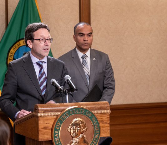 Washington State Reacts to Feared ICE Invasion, Constitutional Crisis Ferguson stands at the lectern with Brown over his shoulder. Both wear suits and ties.