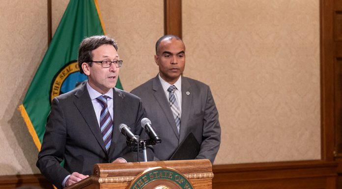Washington State Reacts to Feared ICE Invasion, Constitutional Crisis Ferguson stands at the lectern with Brown over his shoulder. Both wear suits and ties.
