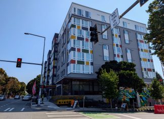 Seattle Leads Nation in Affordable Apartment Production A seven story apartment building with gray and white siding with red, gold, and green accents. Photo shows the view from the nearby intersection.