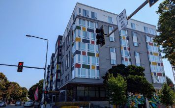 Seattle Leads Nation in Affordable Apartment Production A seven story apartment building with gray and white siding with red, gold, and green accents. Photo shows the view from the nearby intersection.