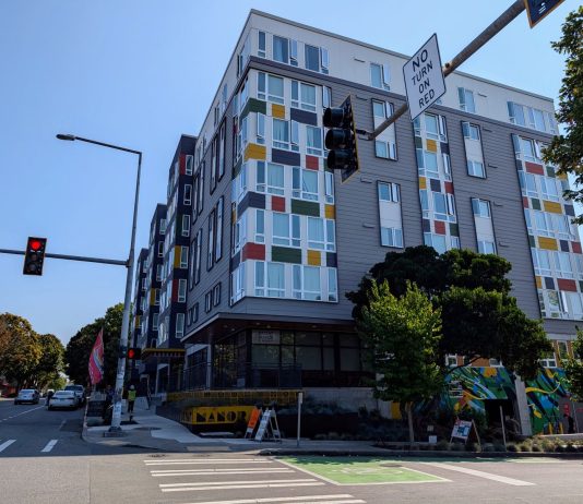 Seattle Leads Nation in Affordable Apartment Production A seven story apartment building with gray and white siding with red, gold, and green accents. Photo shows the view from the nearby intersection.