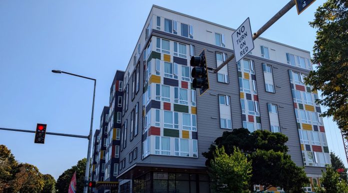 Seattle Leads Nation in Affordable Apartment Production A seven story apartment building with gray and white siding with red, gold, and green accents. Photo shows the view from the nearby intersection.