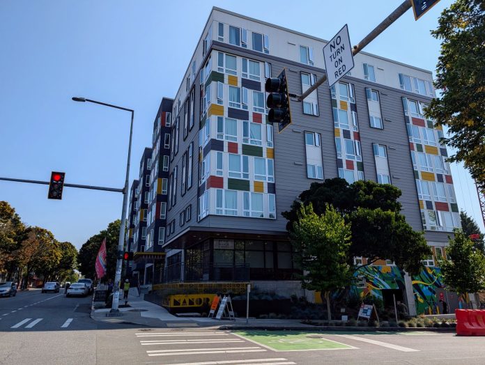 A seven story apartment building with gray and white siding with red, gold, and green accents. Photo shows the view from the nearby intersection.