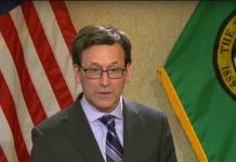 Ferguson’s 2026 Budget Queues Steep Cuts, Pushes Millionaires Tax to 2029 Ferguson wears a dark gray suit and tie and stands in front of the flags of the United State and Washington state.