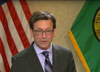 Ferguson’s 2026 Budget Queues Steep Cuts, Pushes Millionaires Tax to 2029 Ferguson wears a dark gray suit and tie and stands in front of the flags of the United State and Washington state.