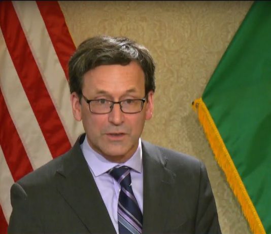 Ferguson’s 2026 Budget Queues Steep Cuts, Pushes Millionaires Tax to 2029 Ferguson wears a dark gray suit and tie and stands in front of the flags of the United State and Washington state.