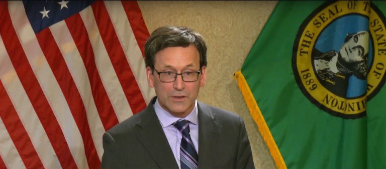 Ferguson wears a dark gray suit and tie and stands in front of the flags of the United State and Washington state.