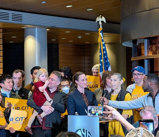 Urbanist Podcast: New Year, New Mayor, New Budget Wilson supporters hold yellow "This is your city" signs. She stands at the lectern and and a man adjust the mic to her height.