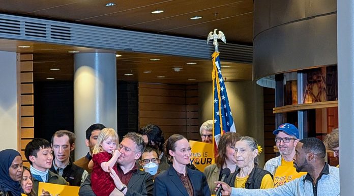 Urbanist Podcast: New Year, New Mayor, New Budget Wilson supporters hold yellow "This is your city" signs. She stands at the lectern and and a man adjust the mic to her height.