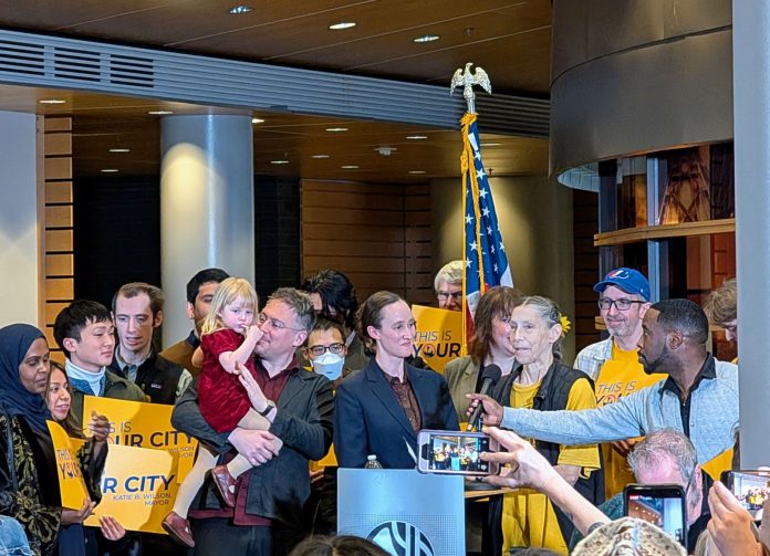 Wilson supporters hold yellow "This is your city" signs. She stands at the lectern and and a man adjust the mic to her height.