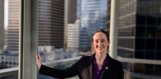 Wilson wears a black blazer and stands on the balcony outside her seventh floor office, with the downtown skyline in the background