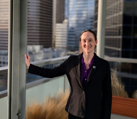 One Week In, Katie Wilson Charts the Path Ahead Wilson wears a black blazer and stands on the balcony outside her seventh floor office, with the downtown skyline in the background
