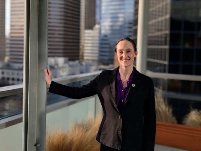 Wilson wears a black blazer and stands on the balcony outside her seventh floor office, with the downtown skyline in the background