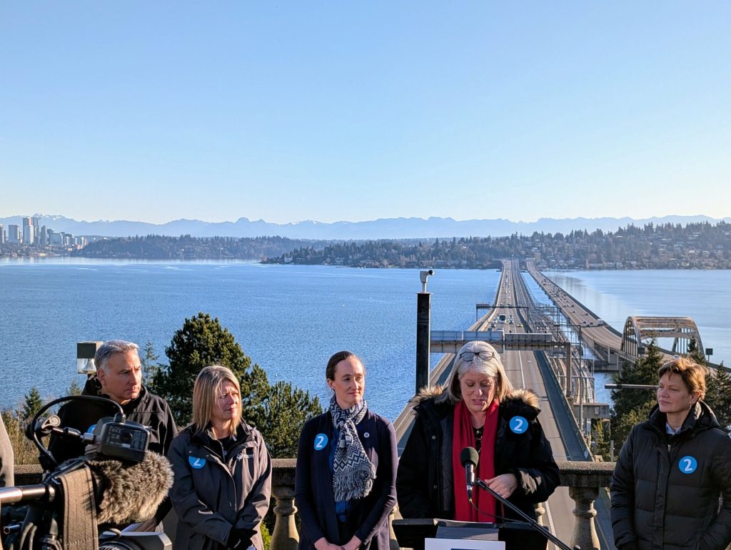Lake Washington and the I-90 floating bridge is visible in the background behind CEO Dow Constantine, Redmond Mayor Angela Birney, Seattle Mayor Katie Wilson, and Claudia Balducci.