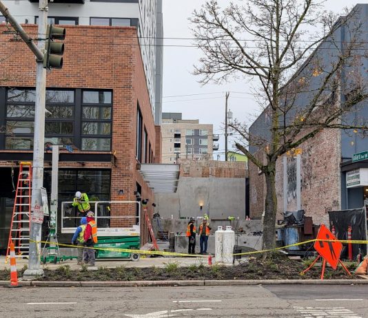 Wilson Promotes Sam Steele to Lead Seattle Permitting Department A half dozen hard-hatted construction workers work at a site between two midrise buildings on Ballard's Market Street.