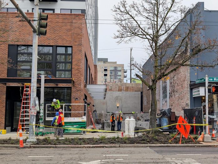 A half dozen hard-hatted construction workers work at a site between two midrise buildings on Ballard's Market Street.