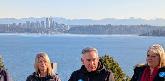From the the I-90 East Portal Viewpoint, Lake Washington and the Downtown Bellevue skyline is visible in the distance. Constantine stands at the lectern gesturing with his hand.