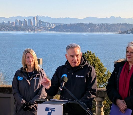 2 Line Countdown Milestone Points to Light Rail Battles Ahead From the the I-90 East Portal Viewpoint, Lake Washington and the Downtown Bellevue skyline is visible in the distance. Constantine stands at the lectern gesturing with his hand.