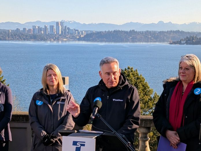 From the the I-90 East Portal Viewpoint, Lake Washington and the Downtown Bellevue skyline is visible in the distance. Constantine stands at the lectern gesturing with his hand.